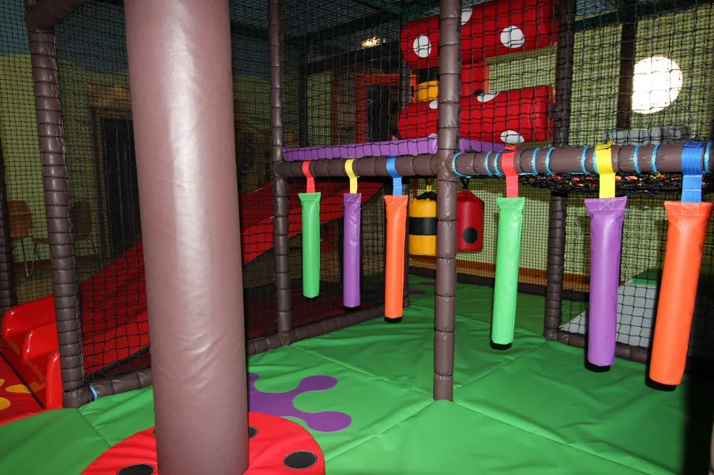 Colourful indoor soft play area with padded poles, netting and foam obstacles on a green floor.