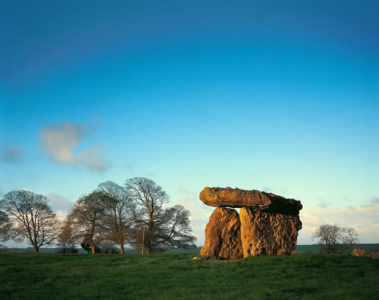 Ancient stone dolmen illuminated by golden sunlight in a grassy field with bare trees under a clear blue sky.