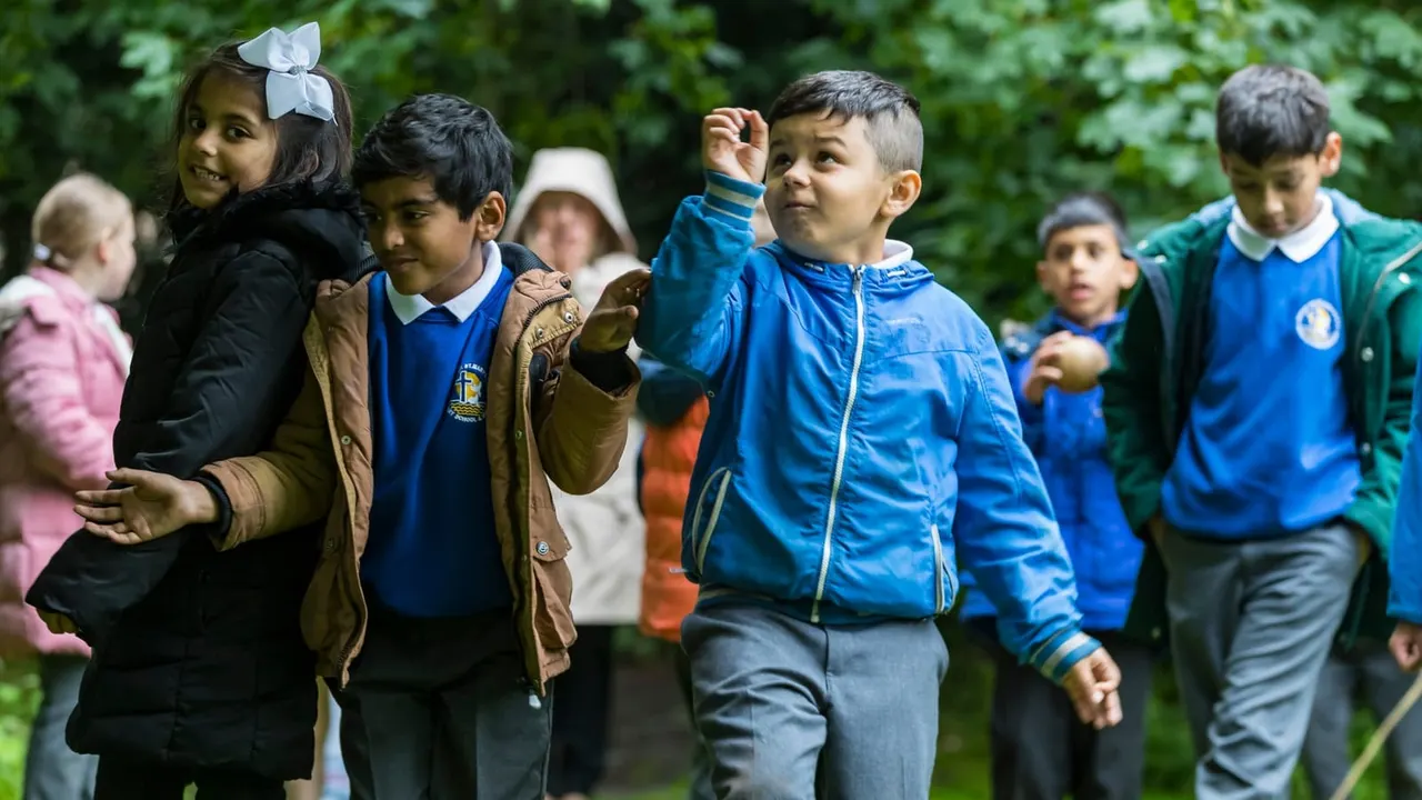 A group of young children walking outdoors amongst green foliage, several wearing blue uniforms and jackets.