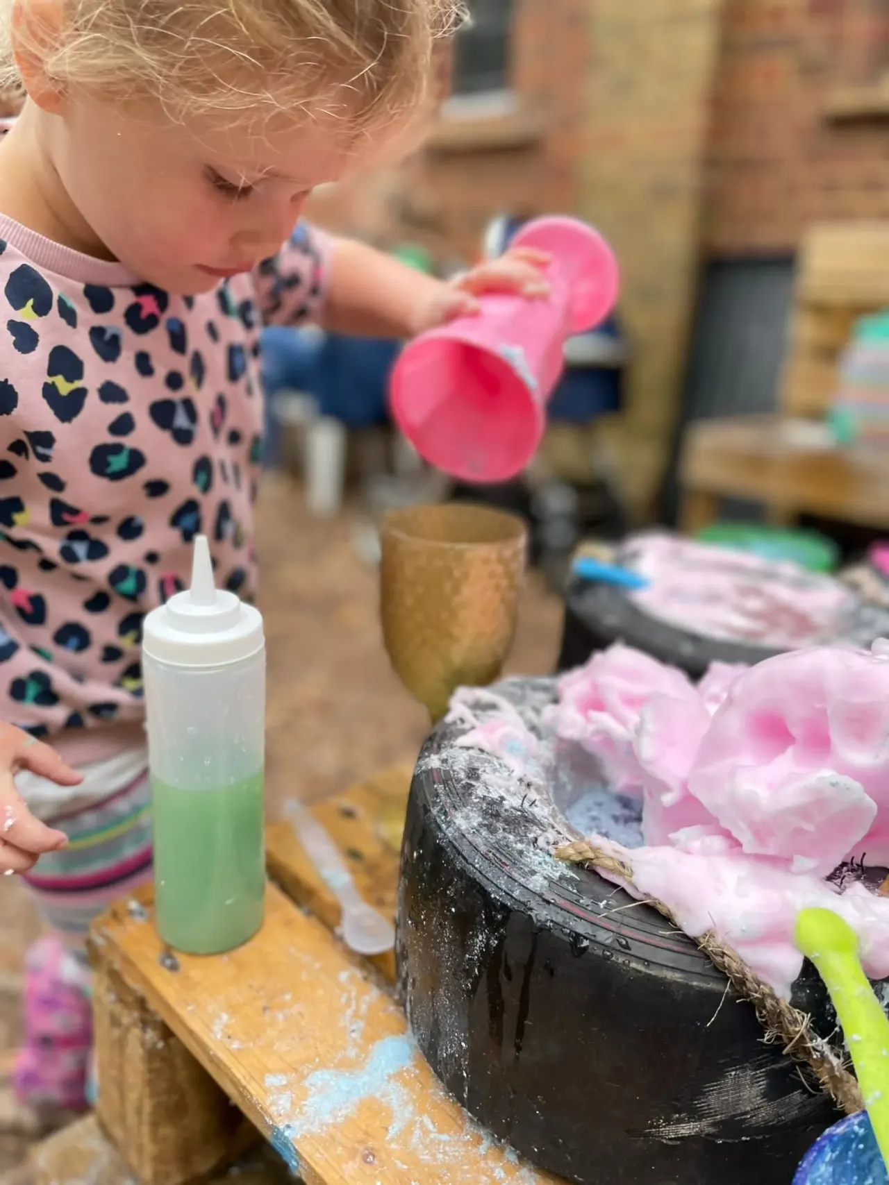 Young child in patterned top engaged in messy play with paint, brushes and materials at an outdoor activity station.