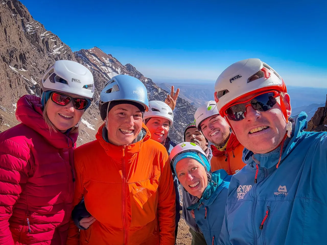 Group of climbers wearing safety helmets and colourful jackets smiling at high altitude on a rocky mountain peak with vast landscape below.