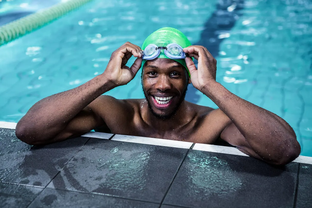 Male swimmer at poolside wearing green swimming cap and goggles, smiling at camera.