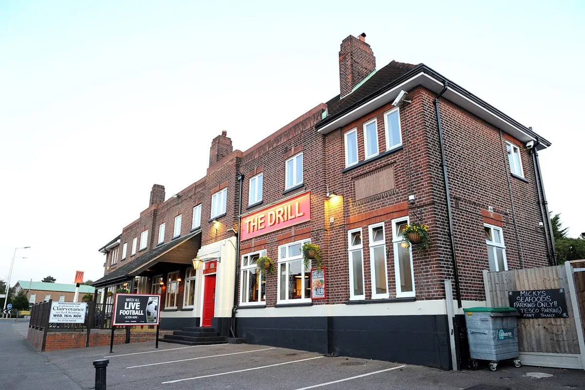 Two-storey brick pub building with "The Drill" signage, multiple chimneys, and hanging baskets, viewed from the street.