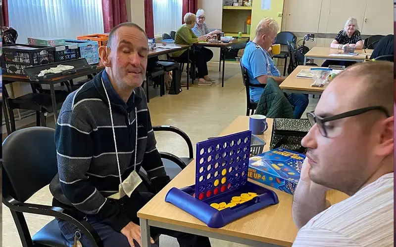 Adults playing Connect Four game at tables in a community centre activity room.