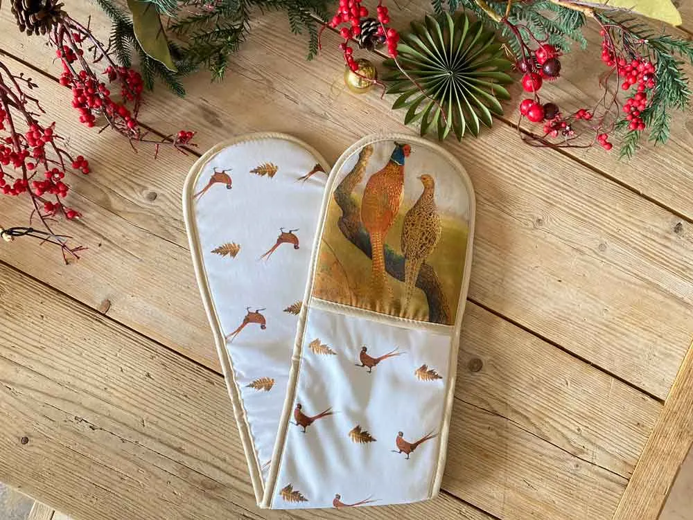 Pair of oven gloves with pheasant and game bird designs displayed on wooden surface with festive foliage.