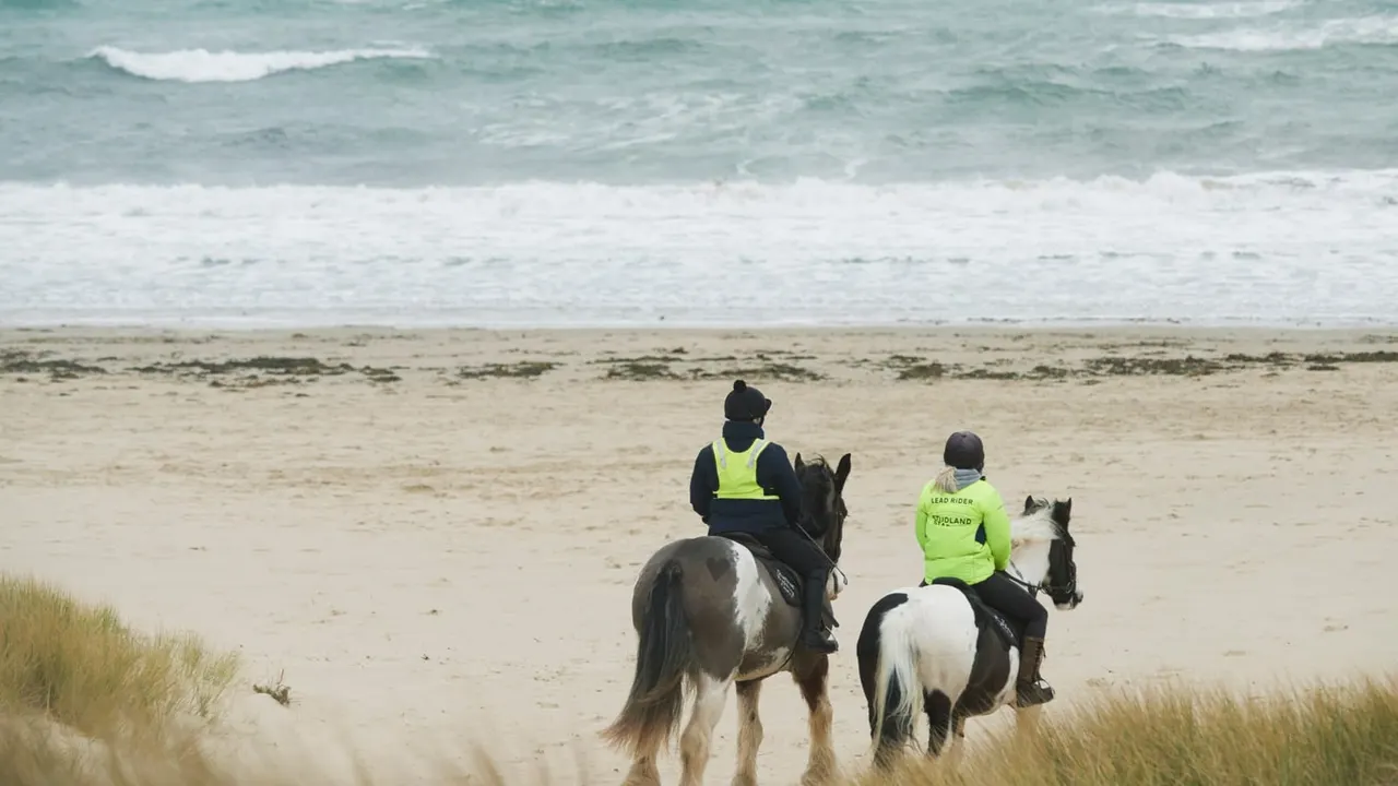 Two riders on horseback wearing high-visibility jackets ride on a sandy beach with waves behind them.