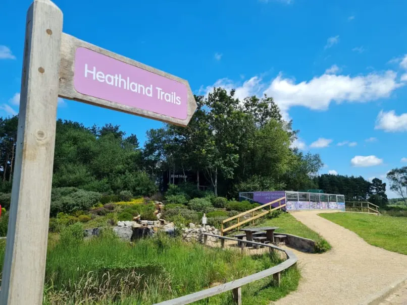 Entrance sign and pathway to Heathland Trails visitor area with wooden structures, landscaped gardens and grassland under blue sky.