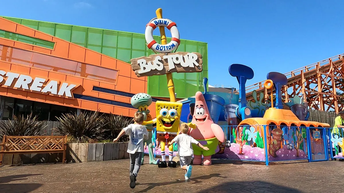 Children running towards large cartoon character mascots in a theme park area with colourful rides and attractions.