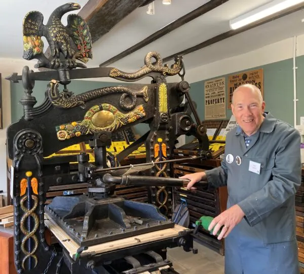 Man in work clothes stands beside an ornate black and gold antique printing press decorated with brass serpents and eagle finials.