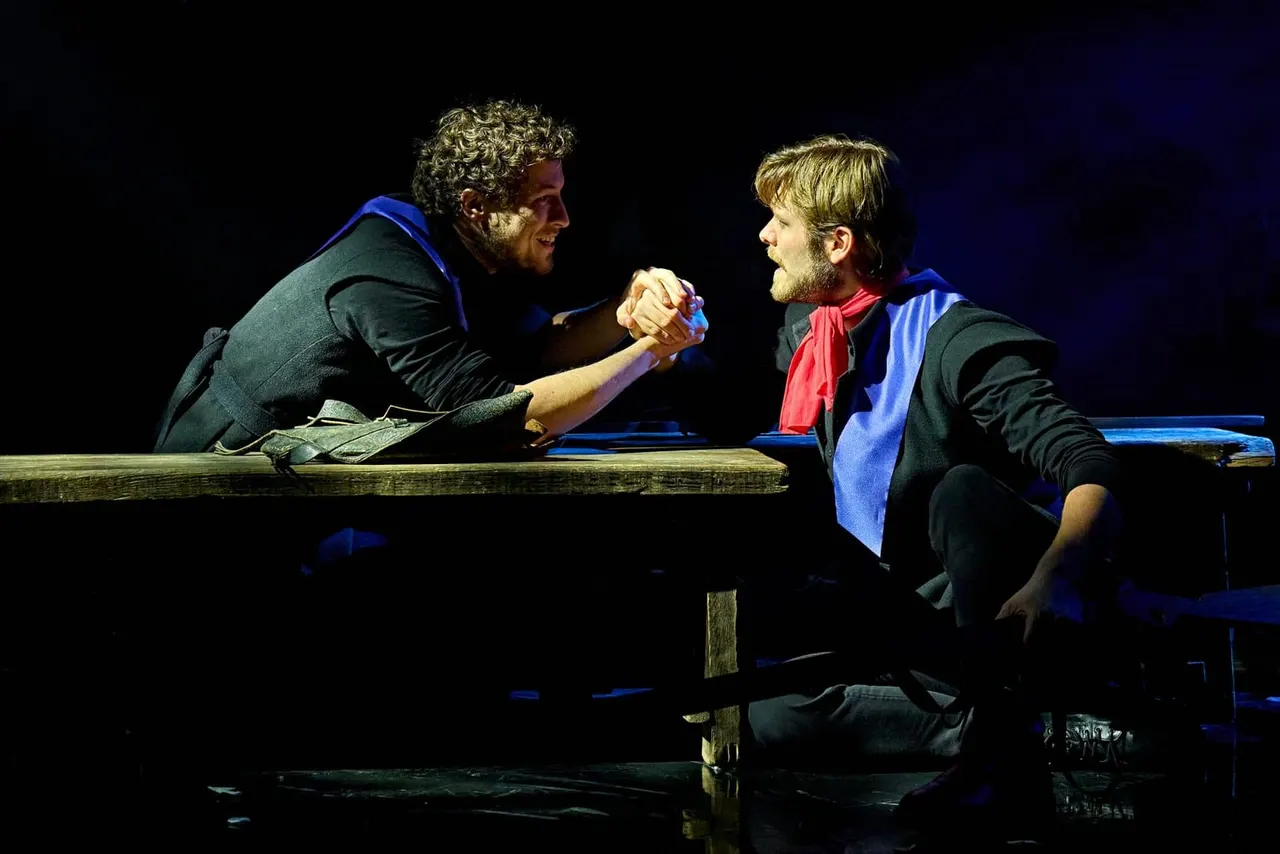 Two men perform a scene on stage, sitting on a wooden platform over water, engaged in an intense conversation under blue lighting.