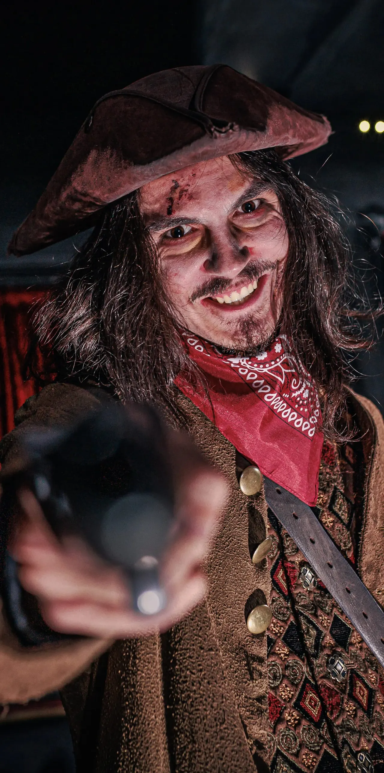 Person in elaborate pirate costume and makeup with tricorn hat and red bandana, smiling at camera in dark indoor setting.