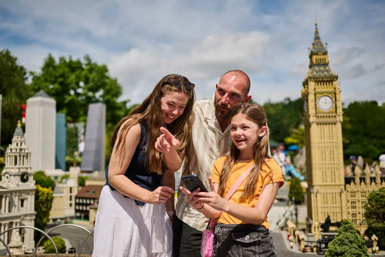 Family of three looking at a phone with miniature landmarks including Big Ben behind them.