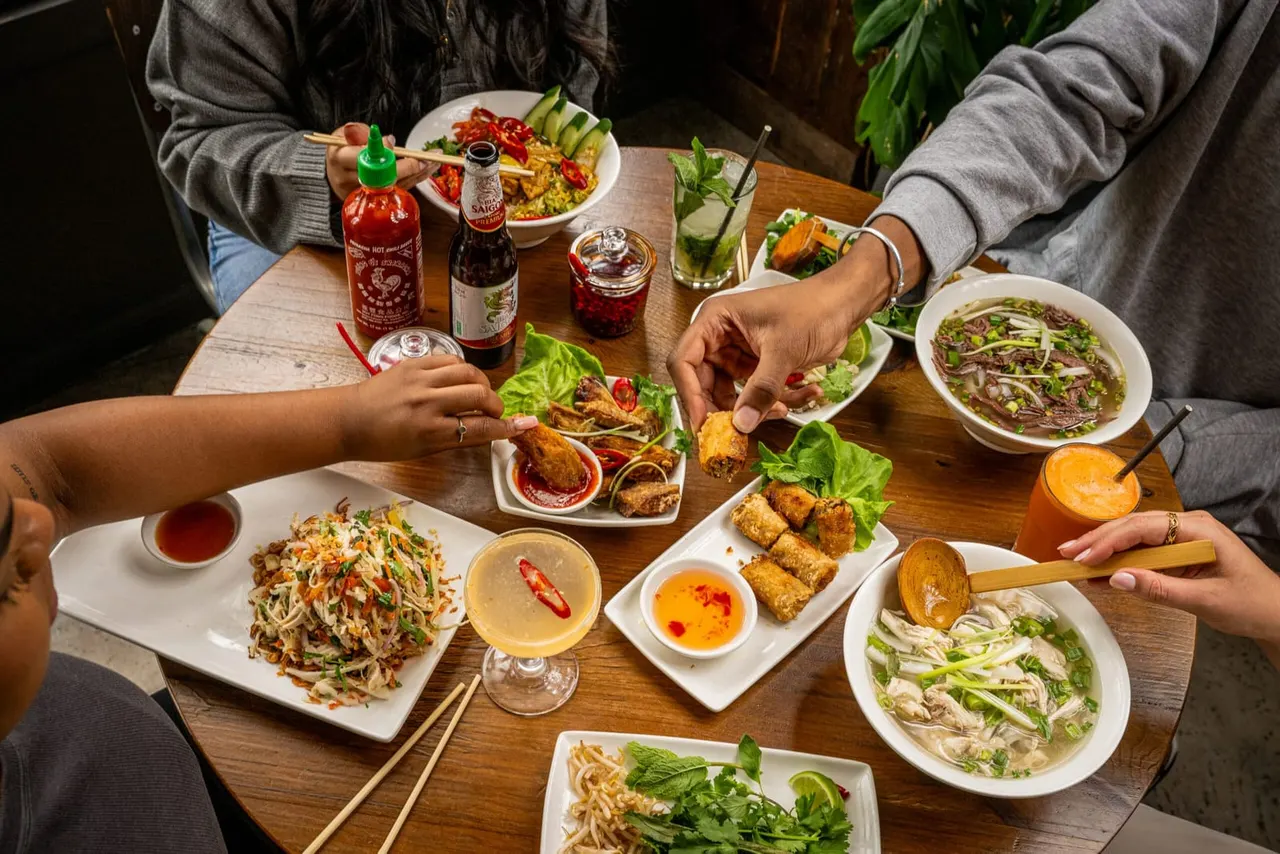 Two people dining together at a wooden table with multiple Asian dishes, drinks and condiments.
