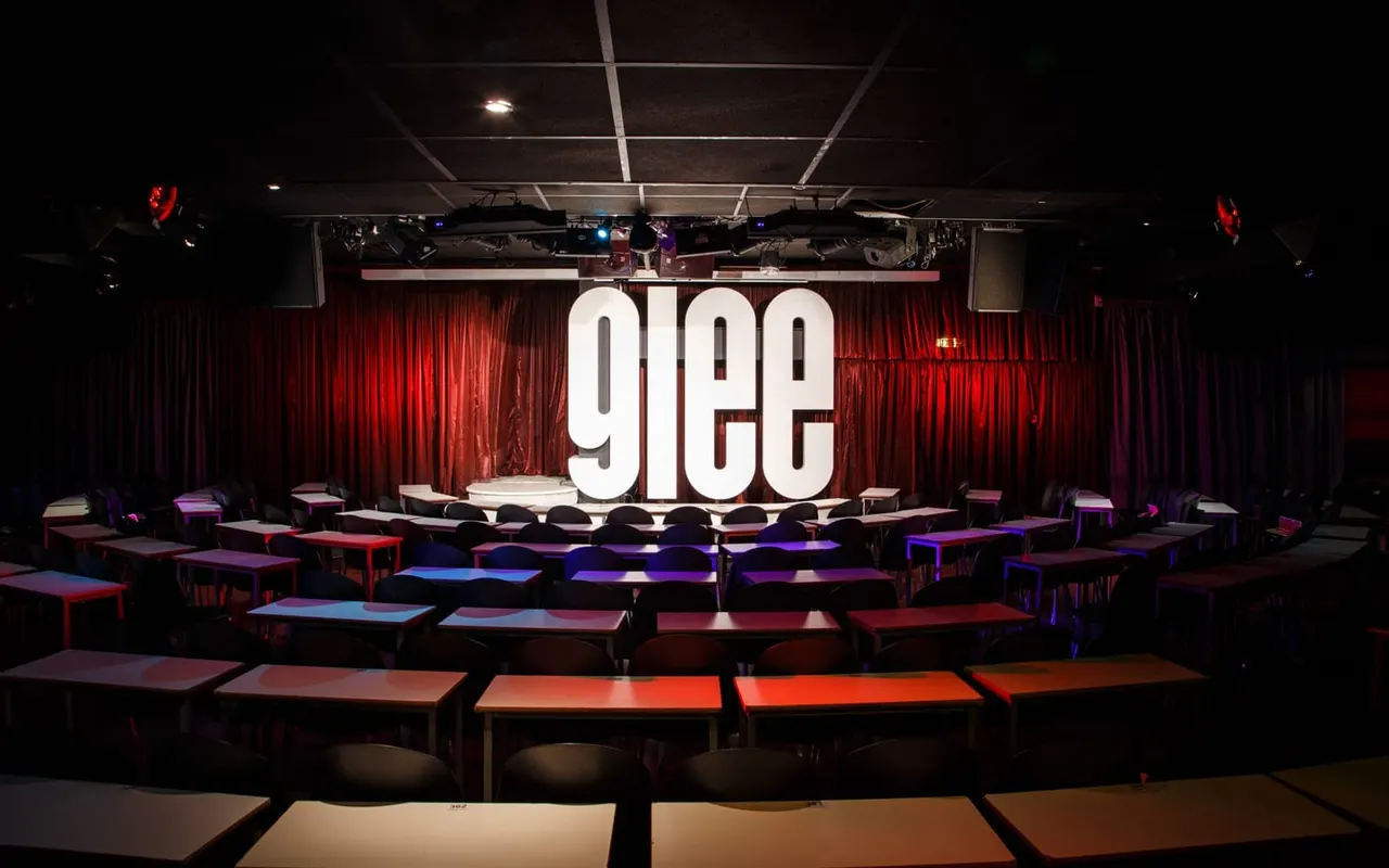 Comedy club interior with illuminated stage, red curtains, colourful seating areas, and professional lighting rig.