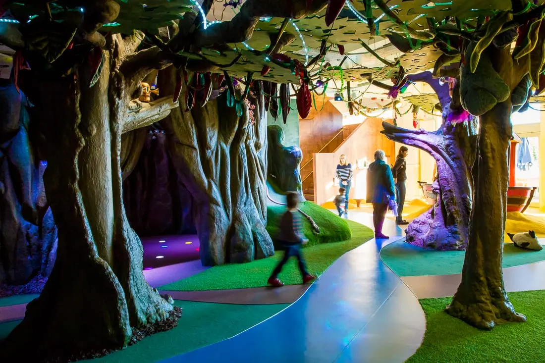 Children and families playing indoor mini golf through a forest-themed course with illuminated tree trunks and coloured lighting.