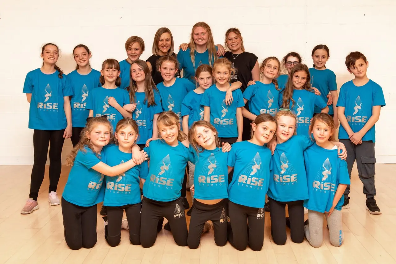 Large group of children wearing matching bright blue Rise Studios t-shirts posing together in a dance studio.