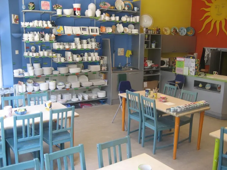 Interior of a ceramics painting studio with blue and wooden chairs at tables, shelves of white ceramic pieces and paints on blue walls.