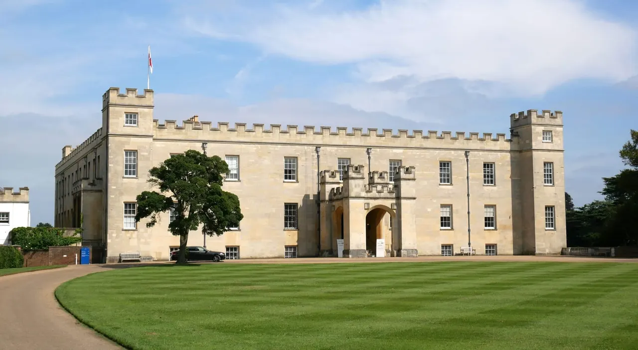 Stone castle with crenellated towers and a large arched entrance, set behind a manicured lawn and tree under blue sky.