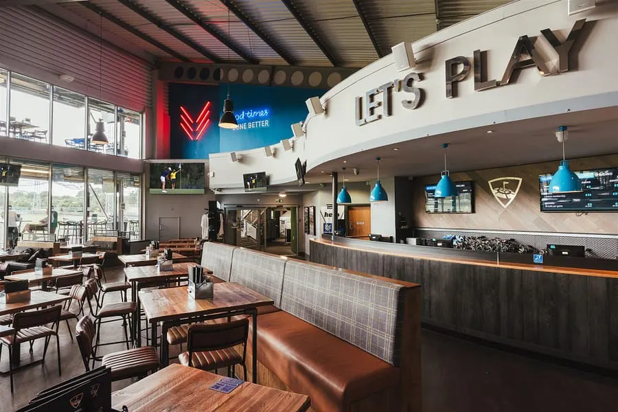Interior of a modern sports leisure venue with dining tables, a service counter, and "Let's Play" signage on the wall.