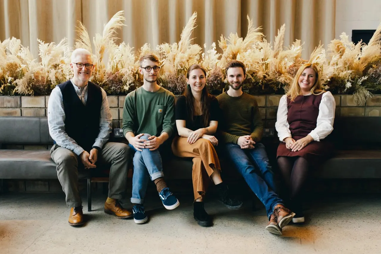Five smiling people seated on a bench against a backdrop of dried pampas grass and neutral curtains.