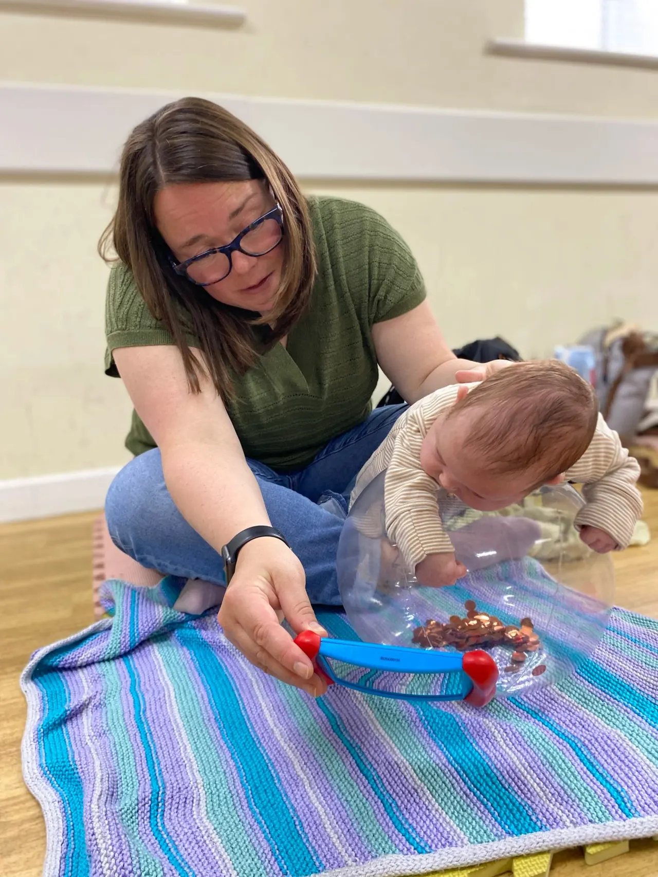 Adult and baby exploring sensory play with tweezers and small objects on a striped mat indoors.
