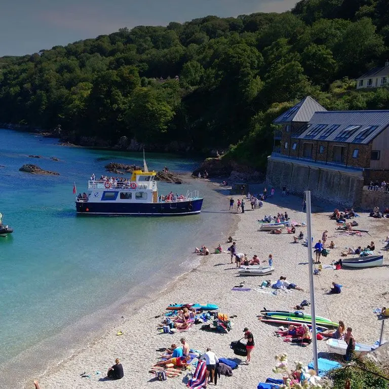 Passenger ferry moored in turquoise water off a pebbly beach lined with sunbathers and boats, surrounded by wooded cliffs.