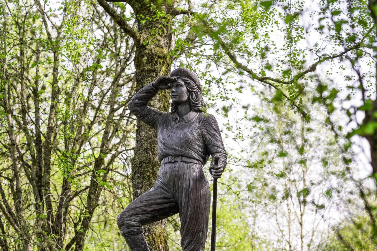 Bronze statue of a female figure in hiking pose amongst woodland trees with spring foliage.