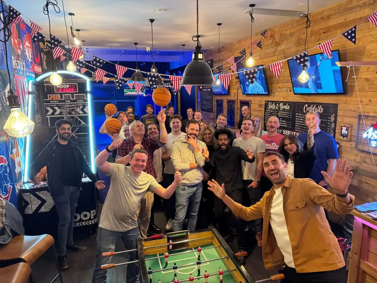 Large group of people enjoying themselves in a sports bar decorated with American flags, basketball hoops and neon signage, with a foosball table in the foreground.