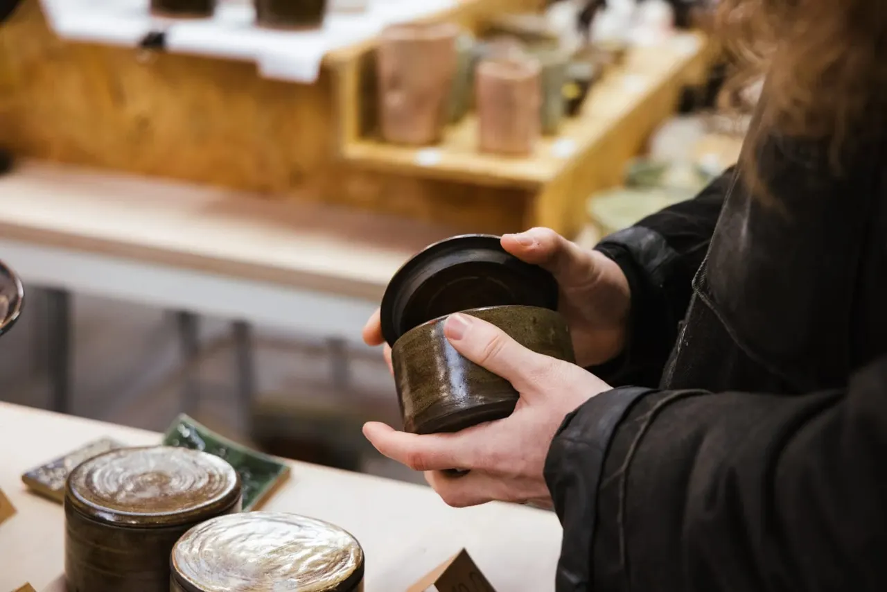 Person in black jacket holding a ceramic pot with lid, examining it at a pottery workshop table.