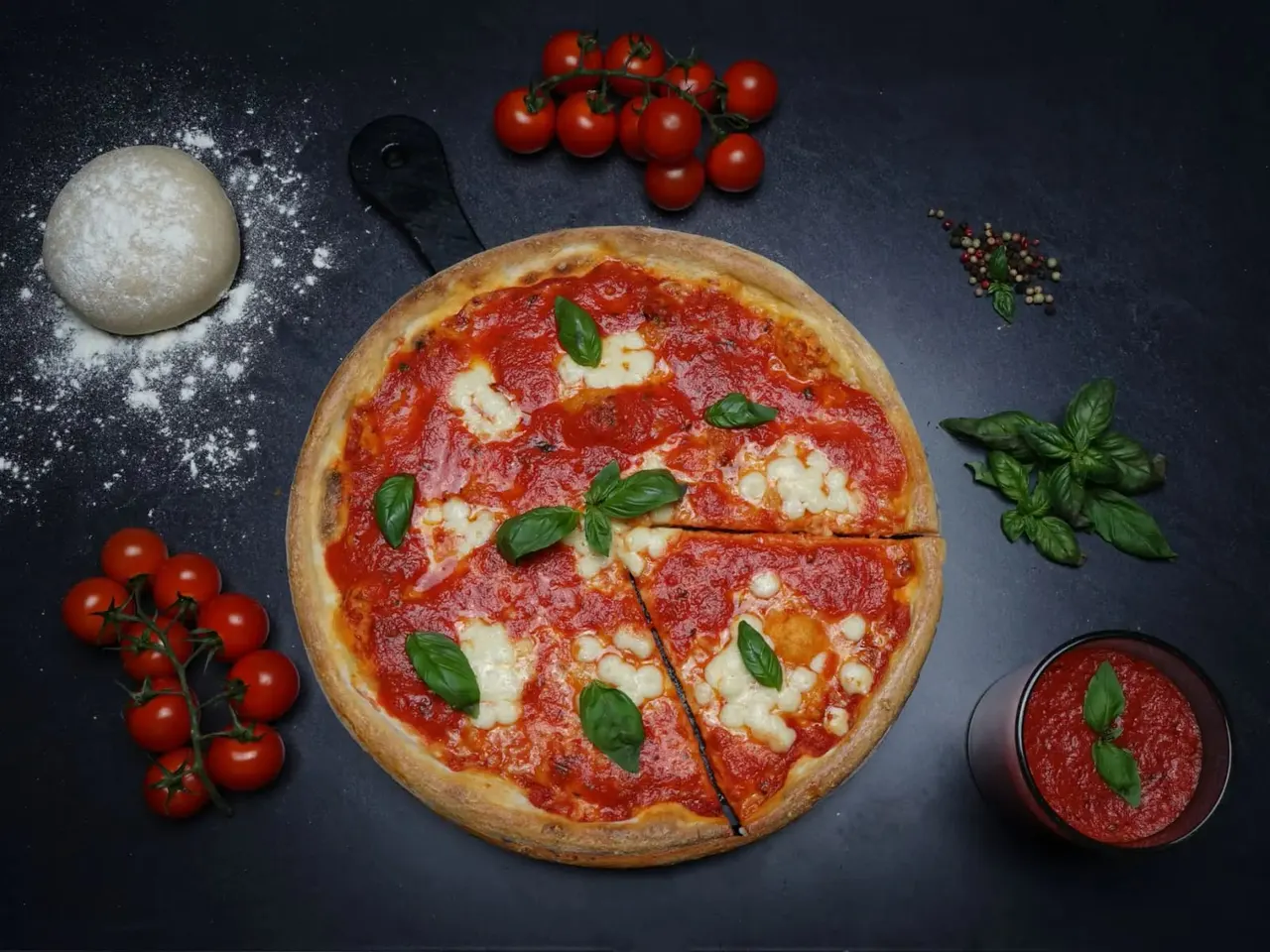 Margherita pizza with tomatoes, basil, flour and ingredients arranged on dark surface.