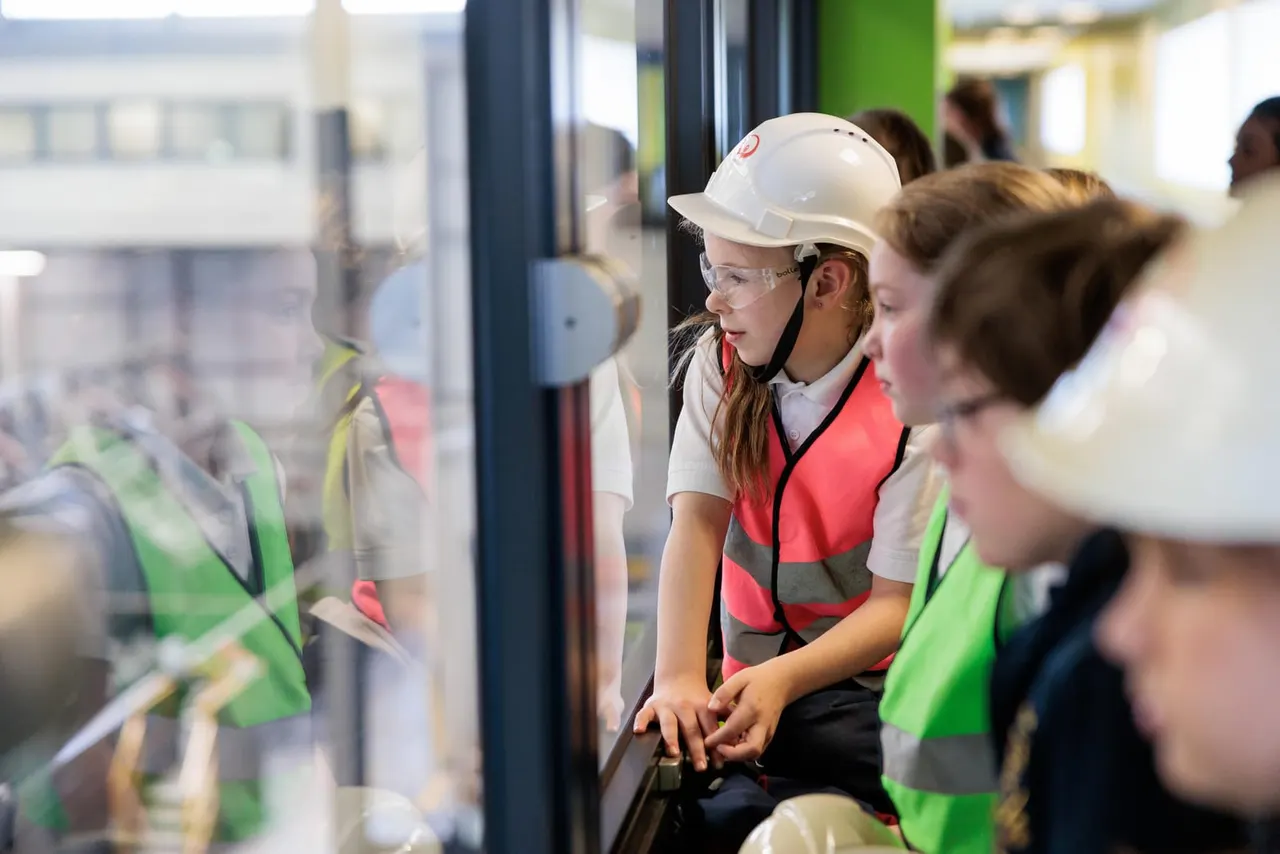 Children wearing hard hats and high-visibility vests observe a recycling facility from an elevated viewing platform.