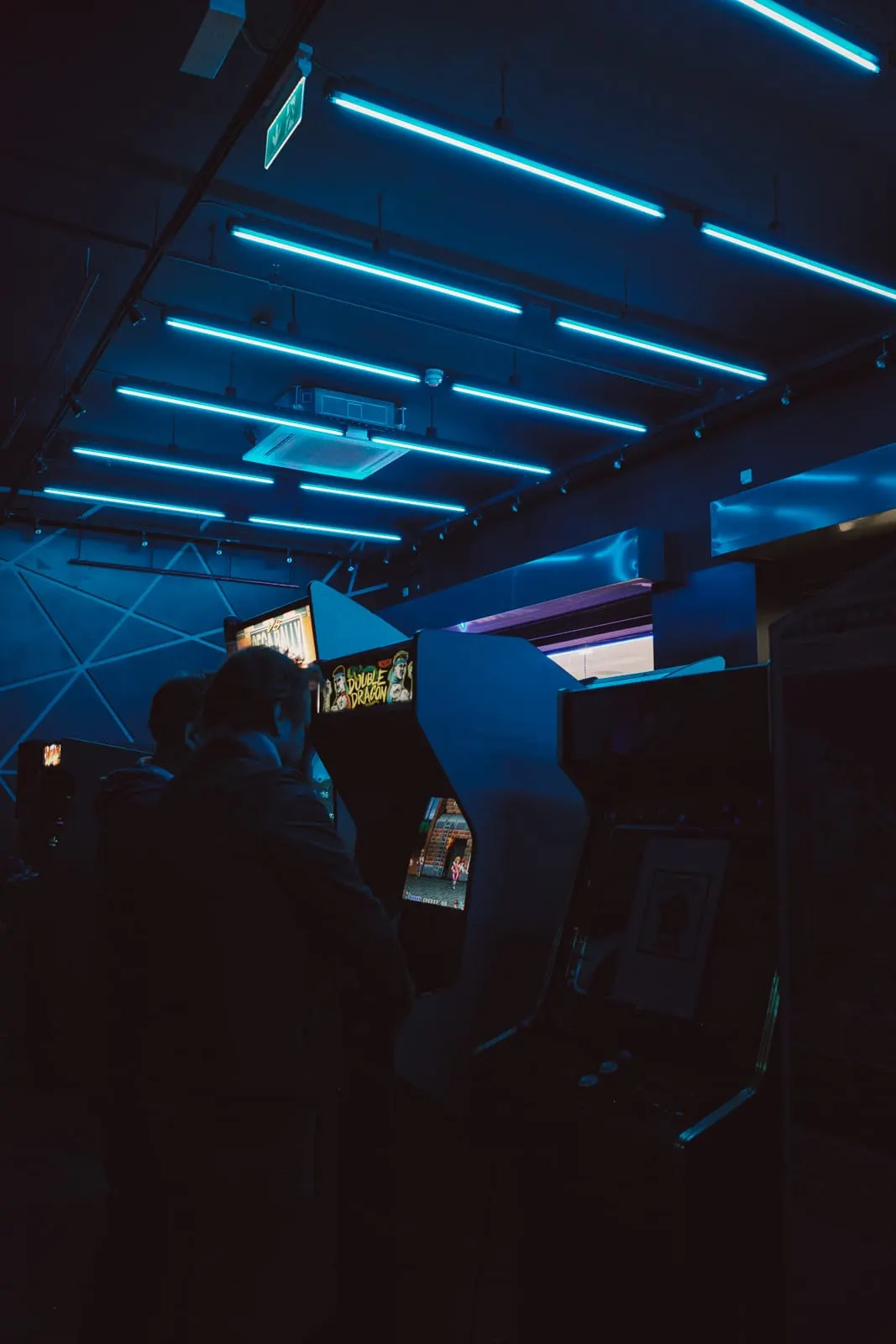 People playing arcade games in a dimly lit venue with bright blue neon lighting overhead.