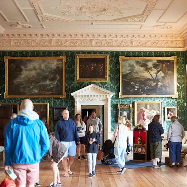 Visitors explore an ornate historic room with painted ceiling, gold-framed paintings on green wallpapered walls, and a white marble fireplace.