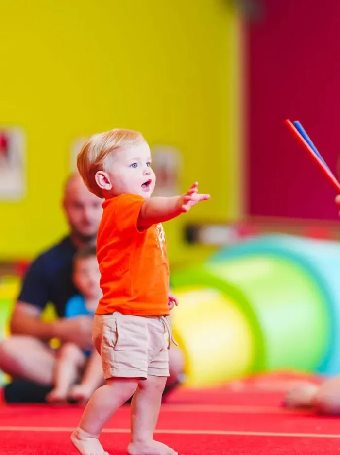 Toddler in orange top playing in a colourful indoor soft play area with foam equipment and bright walls.