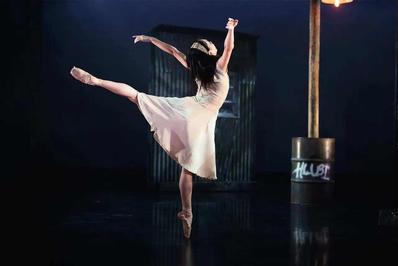 A ballet dancer in a white dress and pointe shoes performs a leap on stage against an industrial backdrop at night.