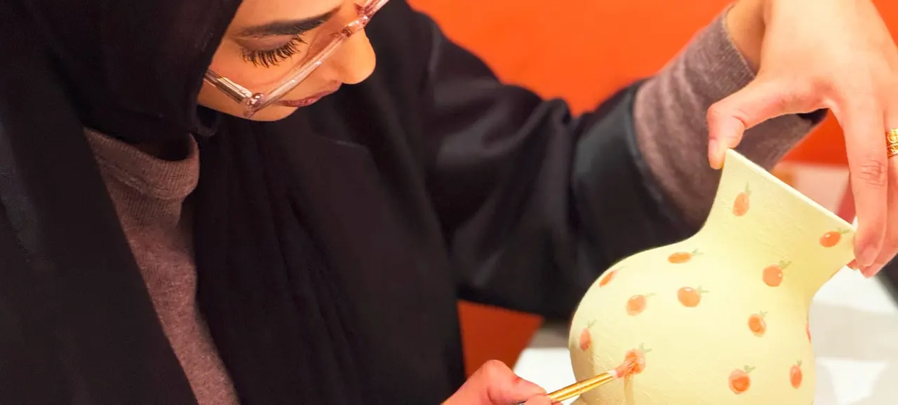 Person painting an orange spotted design on a pale yellow ceramic piece with a fine brush.
