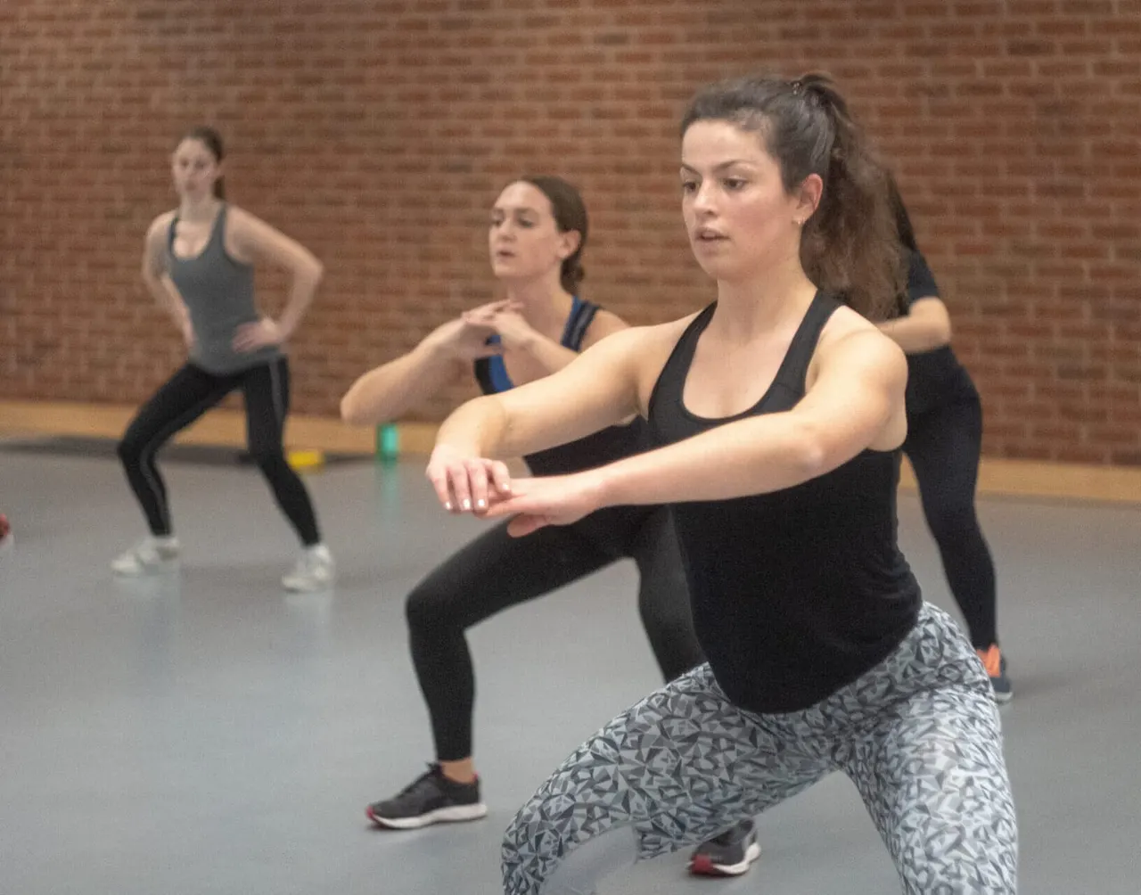 Women participating in a fitness class doing a lunge exercise indoors against a brick wall.
