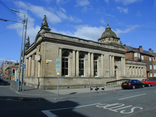 Historic stone library building with domed towers on a street corner with parked cars.