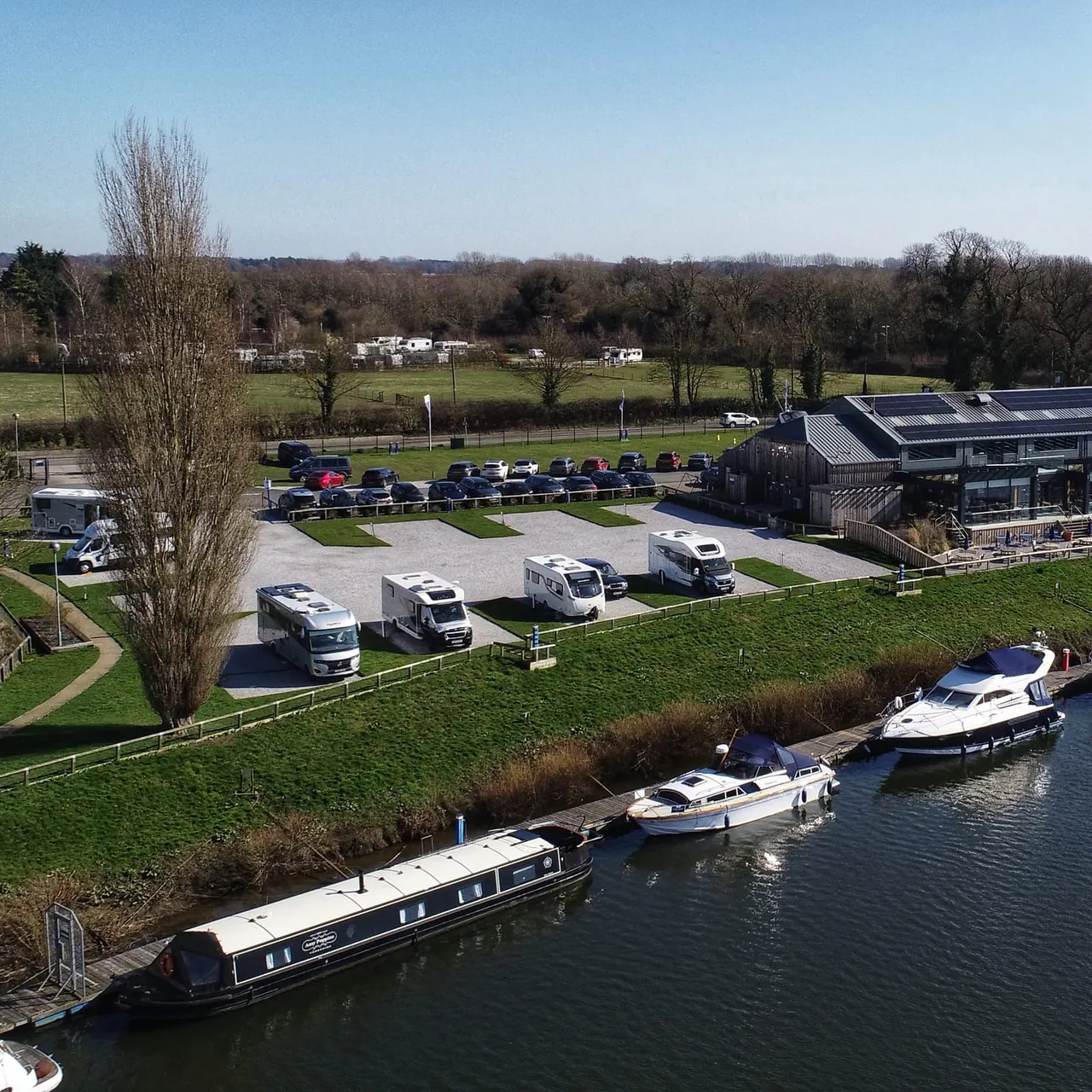 Aerial view of a waterside leisure facility with moored narrowboats, parked caravans and motorhomes, green fields and rural surroundings.