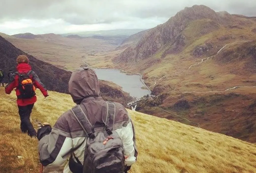 Two hikers with backpacks descending a grassy mountain slope overlooking a lake and valley below.