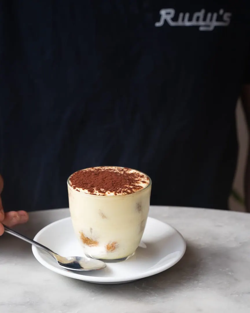 A tiramisu dessert in a glass cup topped with cocoa powder, served on a white plate with a spoon.