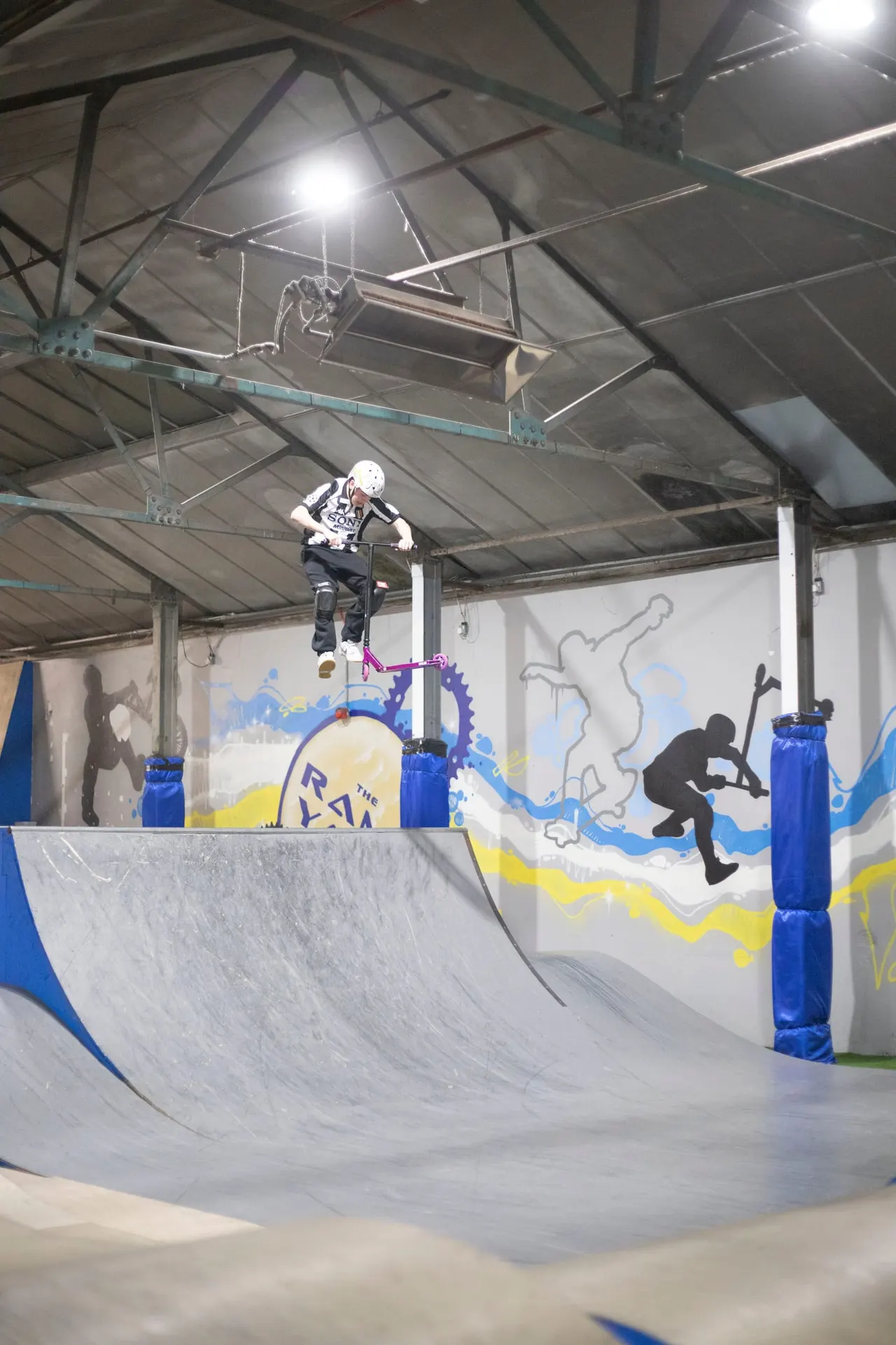 Person in protective gear performing a jump on a scooter at an indoor skate park with ramps and colourful wall graphics.