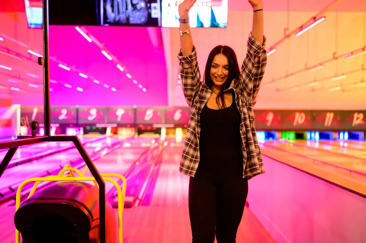 Young woman with arms raised celebrating at a neon-lit bowling alley with colourful lane lighting.