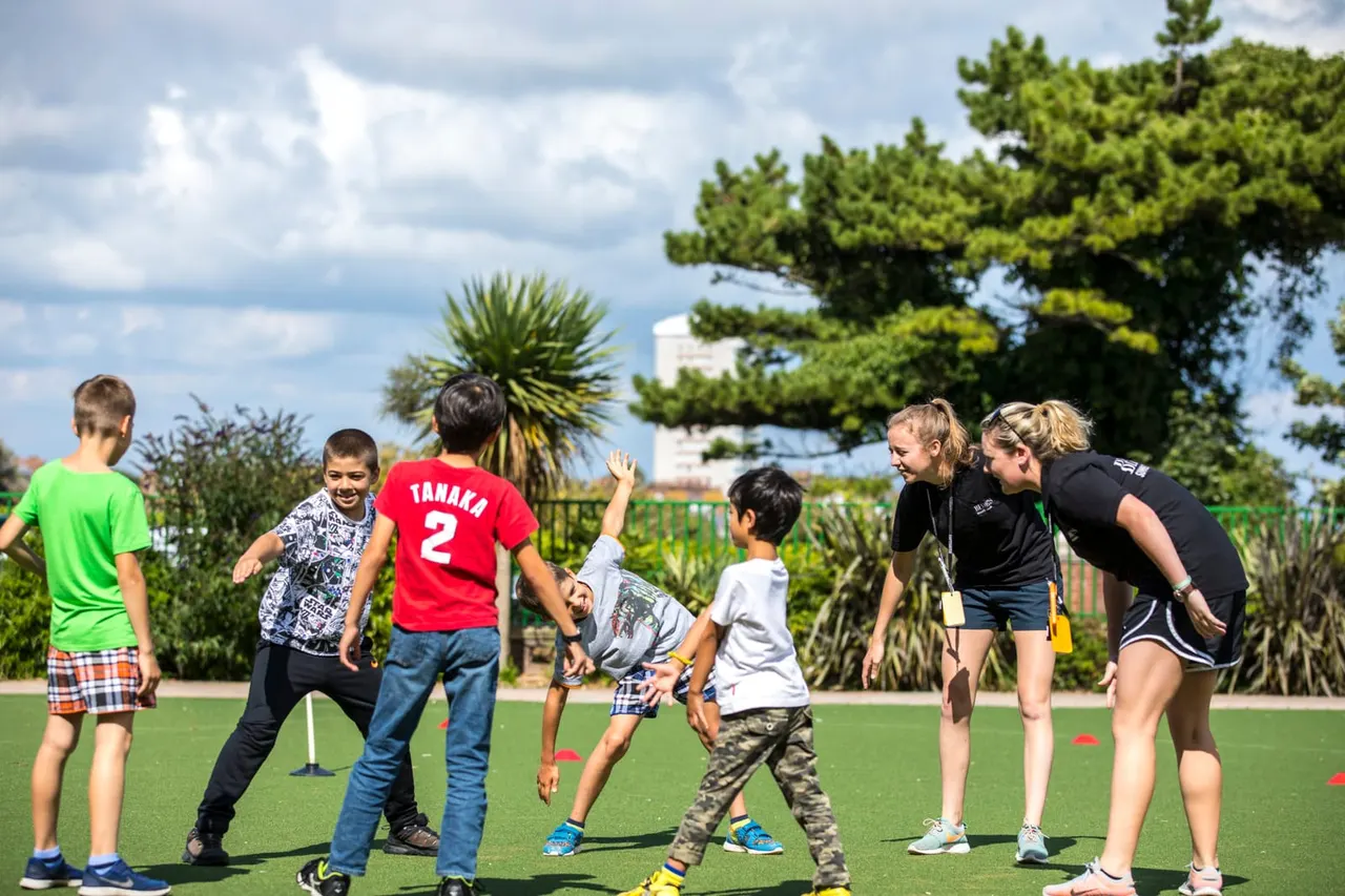 Children playing a sports activity on artificial grass with instructors supervising, overlooking a coastal area with palm trees.