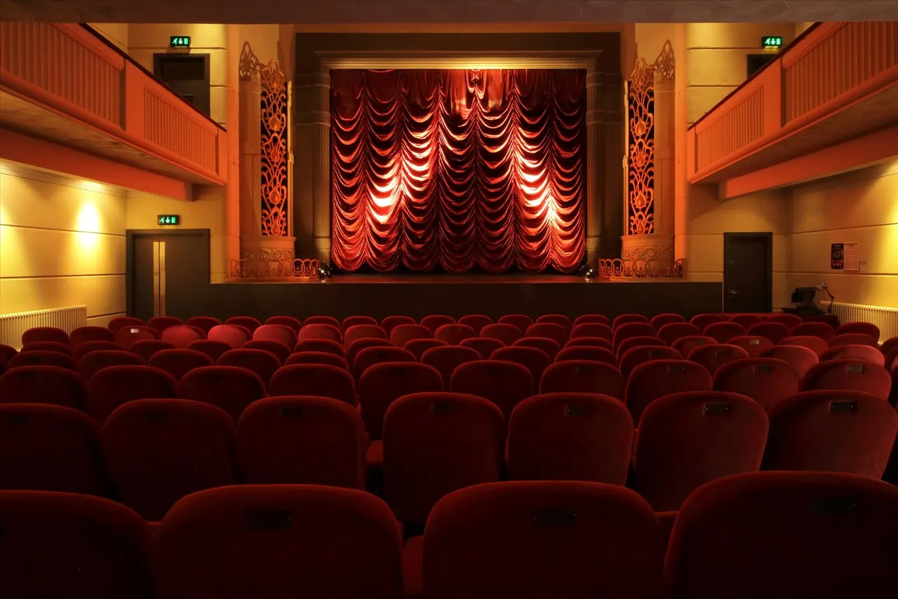 Empty cinema auditorium with rows of red seats facing a stage with dramatic red velvet curtains.