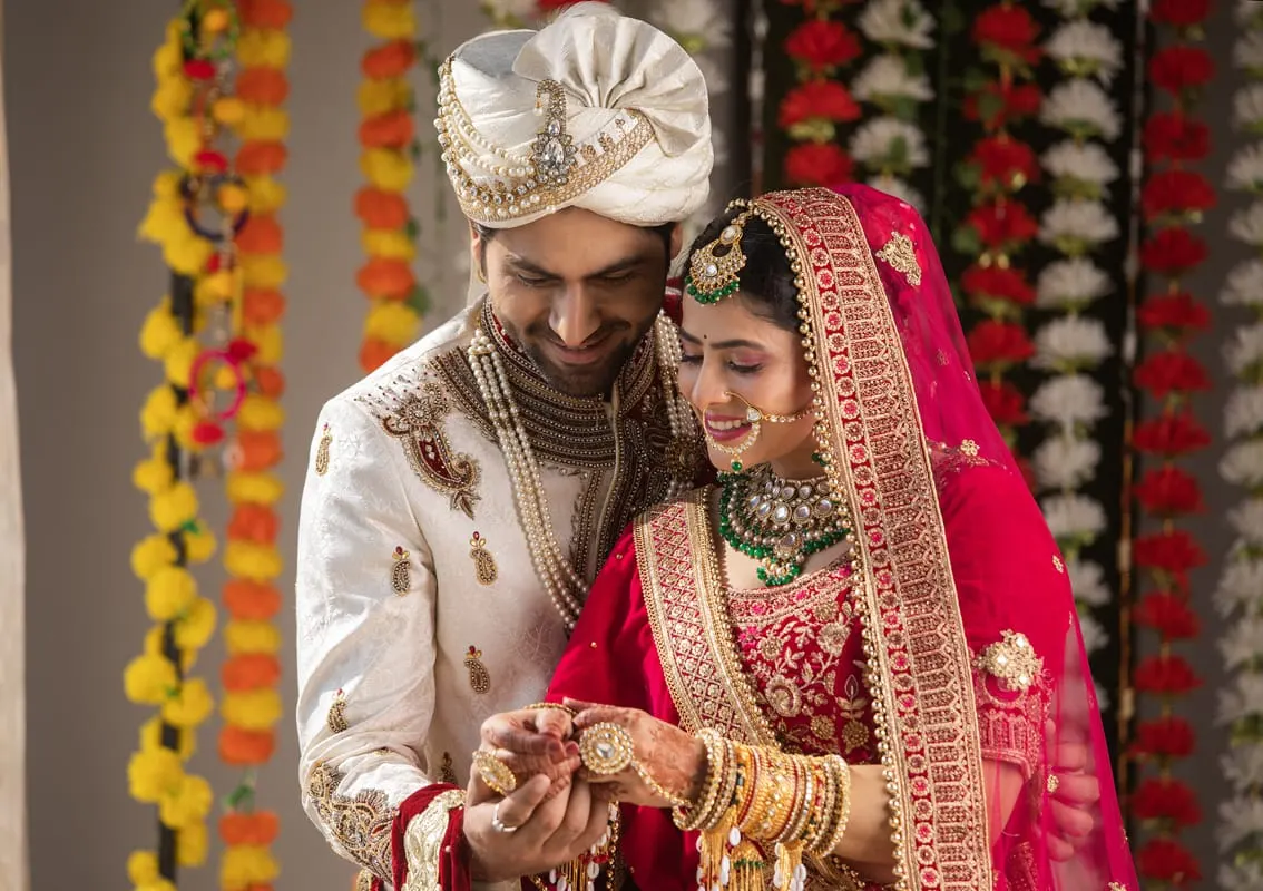 Groom and bride in traditional Indian wedding attire examining a ring, surrounded by floral garlands and decorations.
