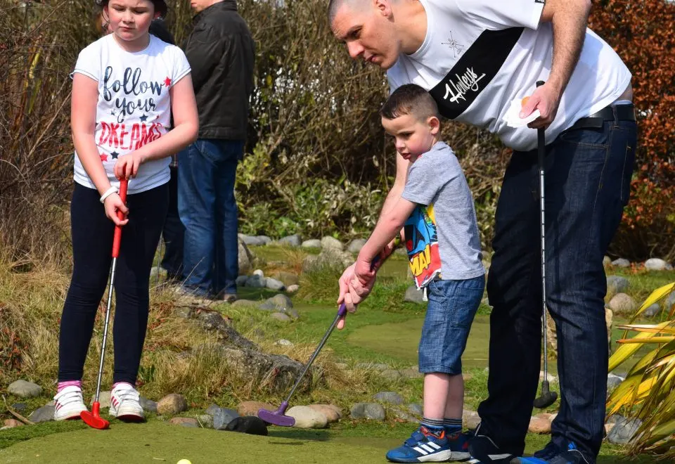 An adult supervises two children playing mini golf outdoors on a green course with shrubs and rocks.