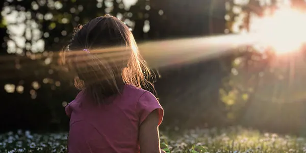 Child in purple shirt sitting on grass at sunset, viewed from behind.