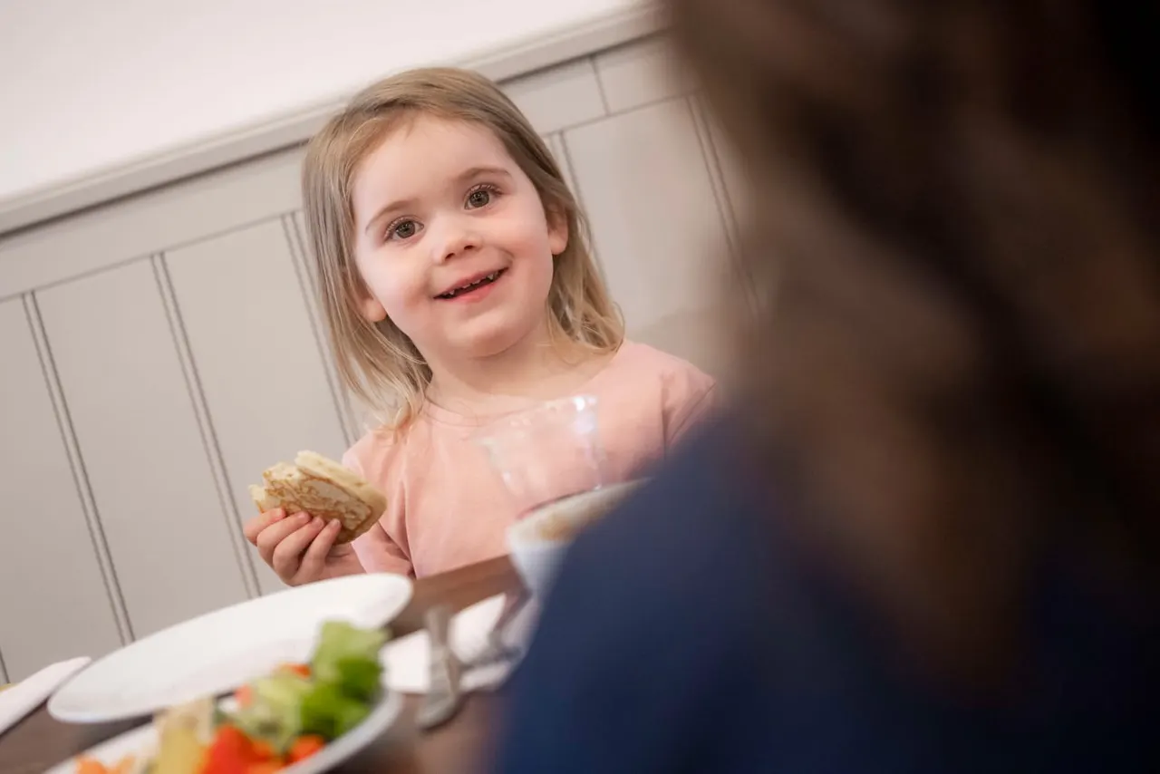 Young child in pink top smiling whilst holding a biscuit at a dining table with food and another person.