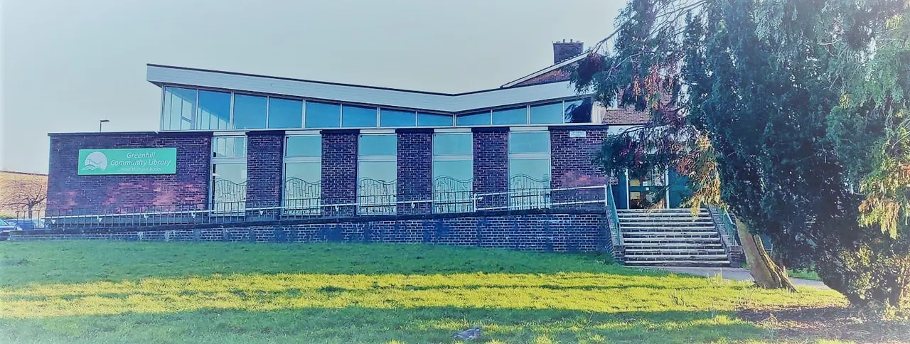 Exterior of a brick community library building with large windows and a sloped modern roof, set on green grass with trees nearby.